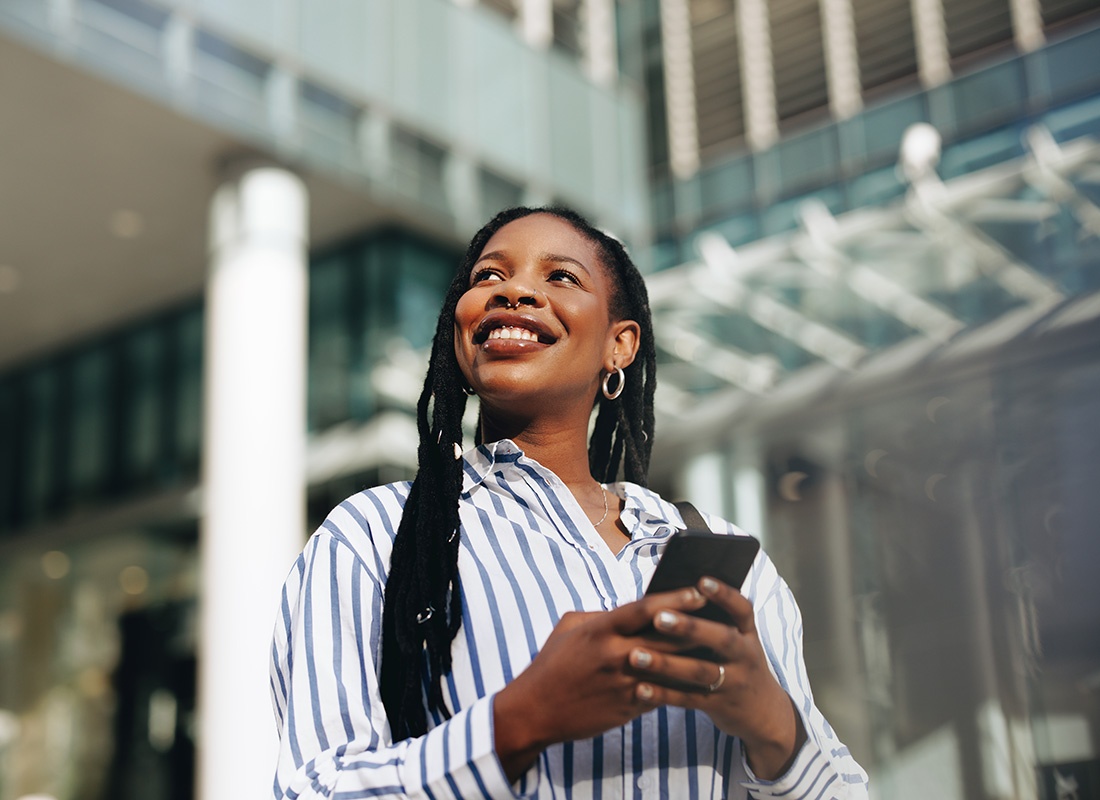 Existing Clients - Portrait of a Smiling Young Business Woman Standing in Front of a Modern Commercial Building on a Sunny Day Whille Holding a Phone in her Hands